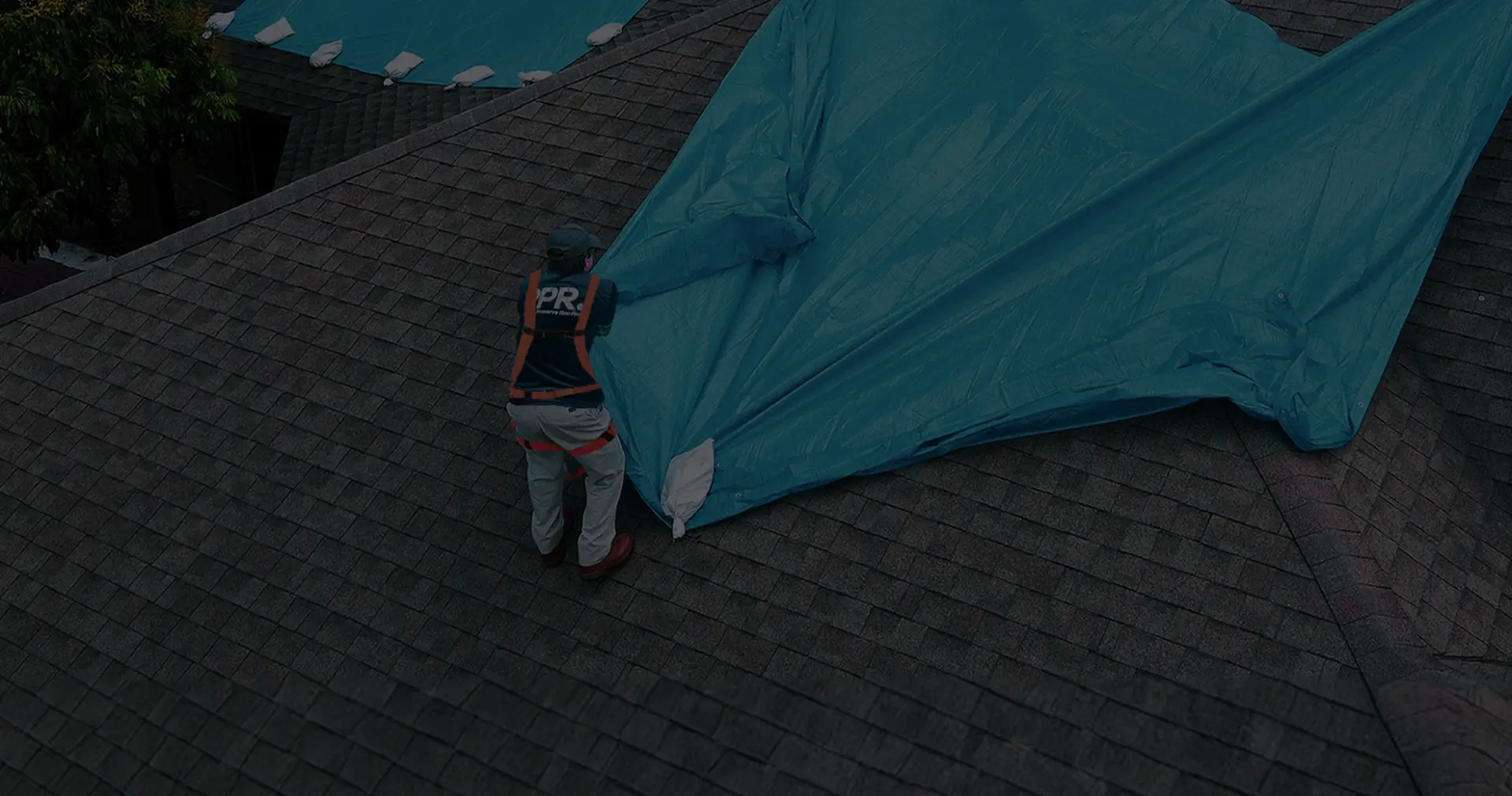 Aerial view of a professional roofer spreading a blue protective tarp over a damaged shingle roof, demonstrating reliable emergency roof tarp service near me.