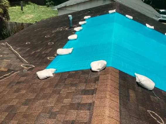 High-angle view of a residential roof with chalk marks identifying storm damage, partially covered by a protective blue tarp from an emergency roof tarp service near me.