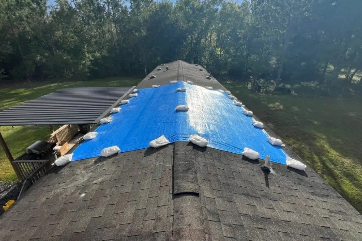 Aerial view of a large residential shingle roof protected by a blue tarp and sandbags, a result of professional emergency roof tarp service near me.