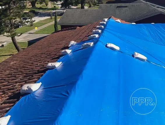 View of a brown tiled roof protected by a blue tarp and white sandbags, illustrating a non-damaging, non penetrating emergency roof tarp service near me.