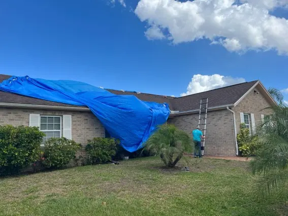 View of a loose, hanging blue tarp on a residential roof with a ladder set up for inspection, highlighting the need for professional emergency roof tarp service near me to properly secure the home.