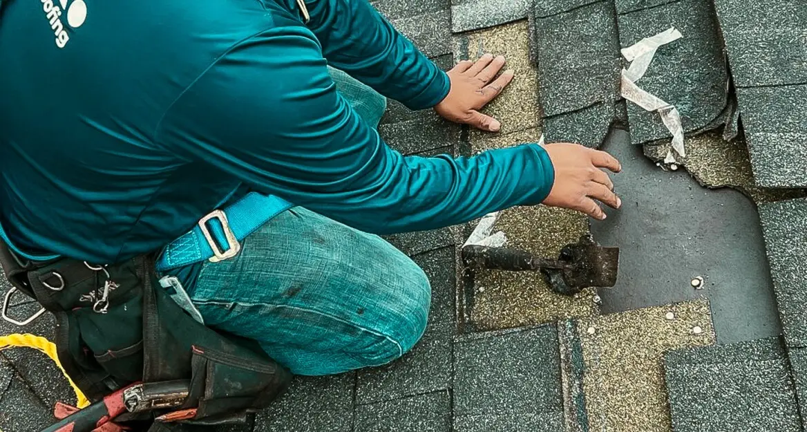 A close-up view of a roofer kneeling on a shingled roof to repair a damaged section. The roofer is wearing a teal long-sleeved shirt, blue jeans, and a safety harness with a tool belt. His hands are near a section of the roof where missing dark shingles expose a yellowish/tan layer and a dark, flat piece of underlayment or flashing. The surrounding shingles are dark gray
