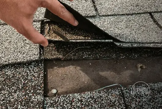 A technician's hand from Protect Preserve Roofing lifting a shingle to identify the source of a roof leak or damage.