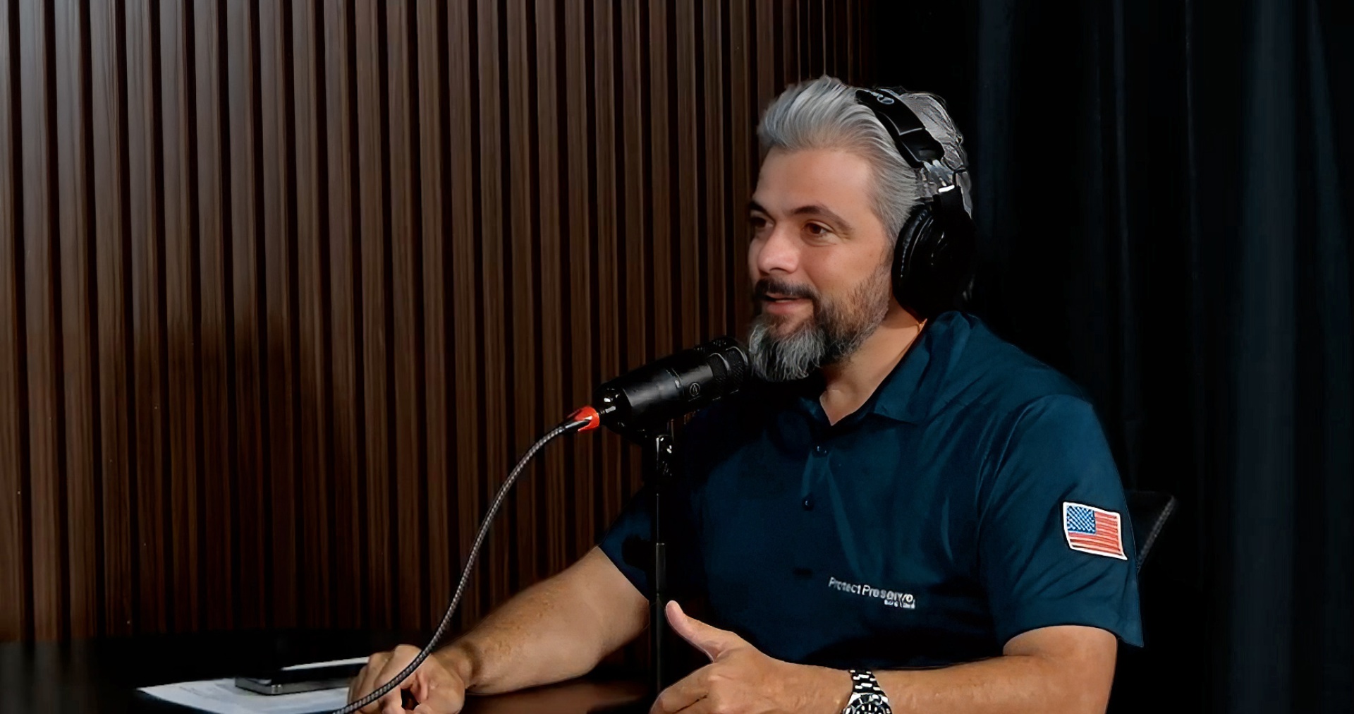 An expert from Protect Preserve Roofing speaking into a microphone during a podcast interview, wearing a branded blue polo shirt.