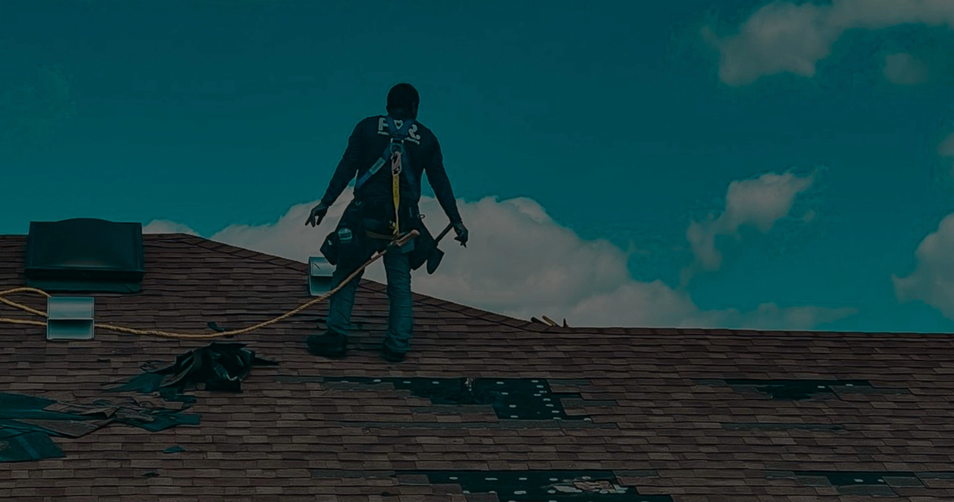 Professional roofer wearing a safety harness standing on a shingle roof while performing expert roof repair services under a blue sky.