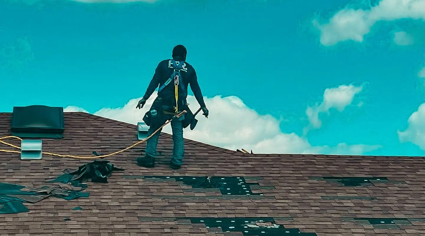 A Protect Preserve Roofing worker repairing a shingle roof under a blue sky with clouds.