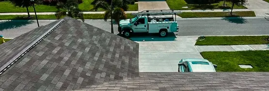 An aerial view looking down onto the gray and dark shingled roof of a house, which dominates the foreground. In the background, a white service truck with ladders on its rack and company lettering on the side is parked on the street next to the driveway. A smaller silver vehicle is partially visible, parked in the driveway below the roofline. The scene includes green lawns and palm trees typical of a suburban neighborhood.