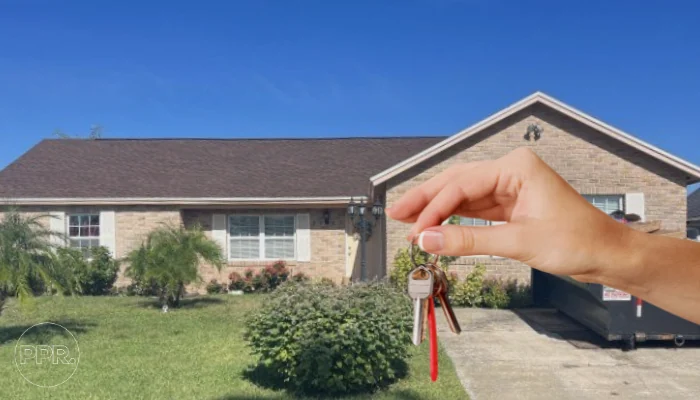 House in the distance with a hand showing keys nearby to represent the keyword asphalt shingles.