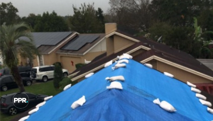 A blue heavy-duty tarp secured over the ridge of a residential roof with sandbags during overcast weather, demonstrating what is the best tarp for a roof to prevent immediate water intrusion.