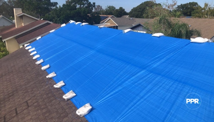 A close-up of a high-quality blue tarp tightly fastened along the edge of a roof with numerous sandbags, illustrating what is the best tarp for a roof to ensure it remains wind-resistant and leak-proof.